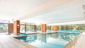 Piscina interna con pareti in vetro trasparente, area a bordo piscina azzurra e vista su spazi verdi esterni.