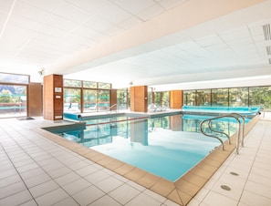 An indoor swimming pool with clear glass walls, a blue poolside area, and a view of greenery outside. - Mountain Flat With Sauna Access (Valloire)
