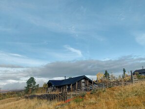 Une cabane rustique, surmontée d’un escalier en bois à marches abruptes, entourée d’une clôture et située au pied d’une colline herbeuse.