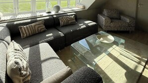 A modern loft bedroom with a large skylight, a sectional sofa, a glass coffee table, and a patterned armchair.