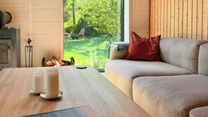 A modern living room with a wooden ceiling, a large window with a view of greenery, a sofa with cushions, and a coffee table with a book and candles. - 60s modernism by Rågeleje beach (Vejby)