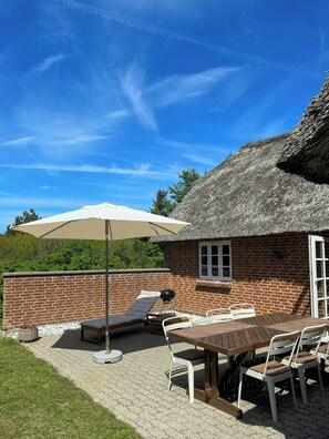 Eine Terrasse mit Tisch und Stühlen, einem Grill und einem Gebäude mit einem Reetdach.