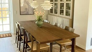 Un espace repas comprenant une longue table en bois, des chaises, des lampes suspendues, un vase avec des fleurs et un meuble avec des portes en verre.