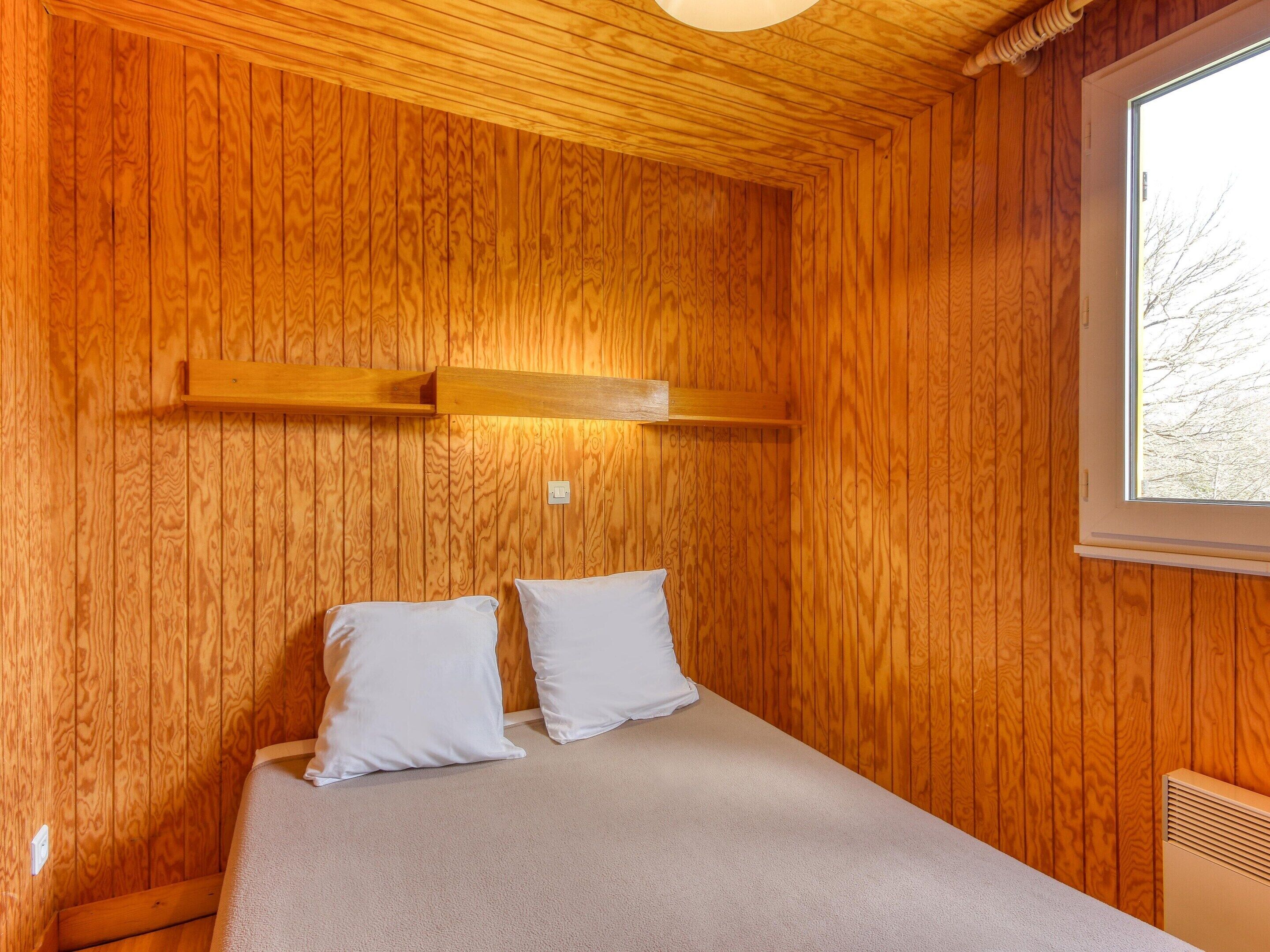 A wooden-paneled room with a bed, a window, and a wall-mounted shelf.