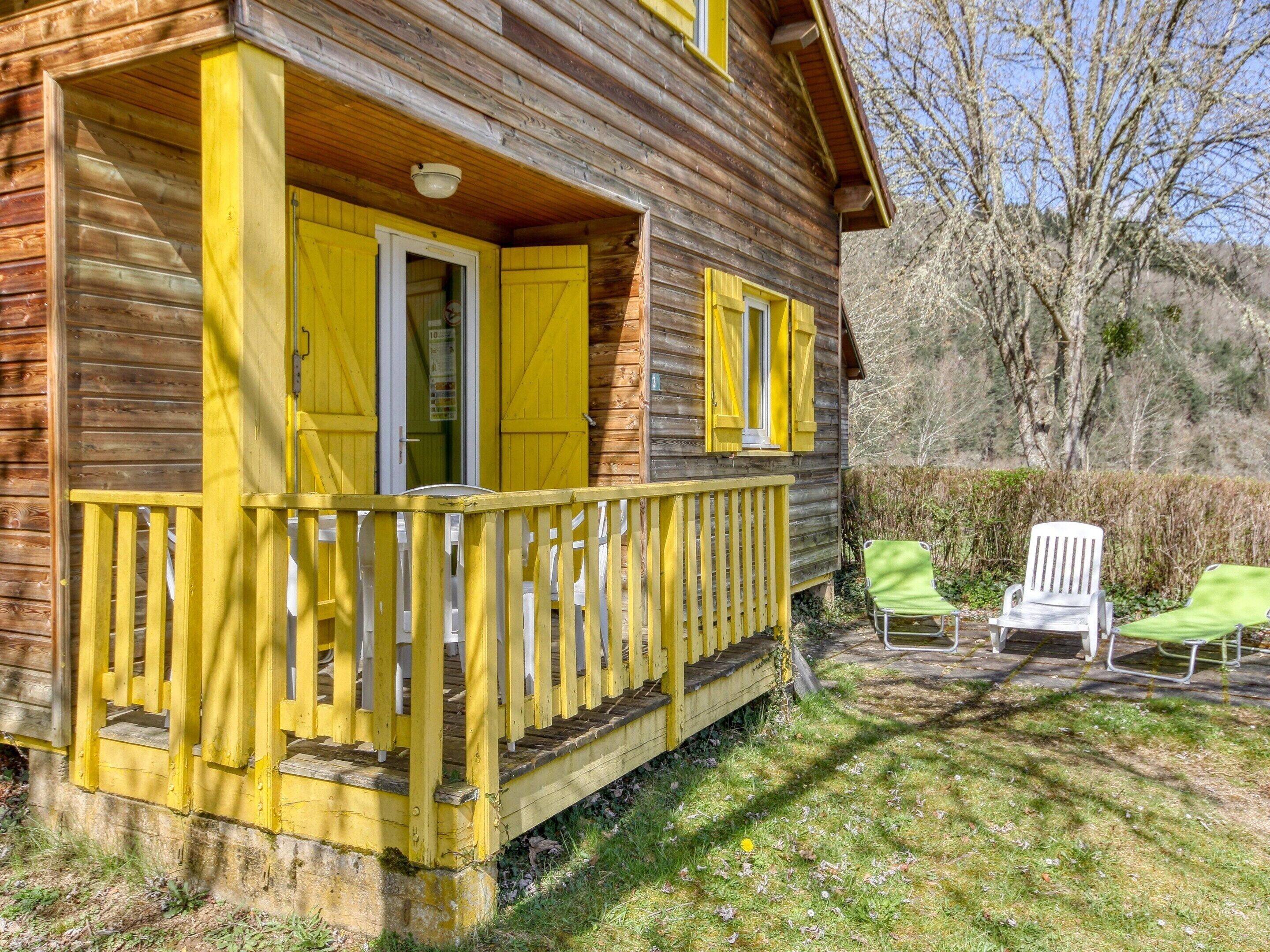 A wooden cabin with yellow shutters, a porch, and two green chairs outside.