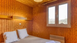 Een kamer met houten lambrisering, een bed, twee witte kussens, een nachtkastje met een handdoek, een raam en een plafondlamp.