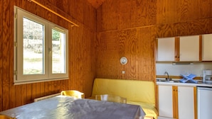 Een houten blokhut van binnen met een eettafel, stoelen, een gele bank, een keuken met kastjes en een gootsteen, en een raam met uitzicht op groen.