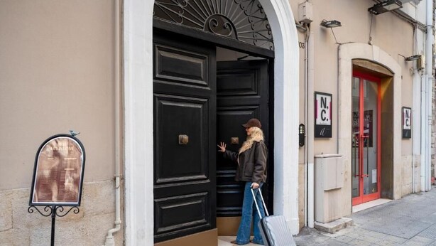 Une personne entre dans un bâtiment qui a une porte noire et une porte rouge, un panneau avec l’inscription « N.C. » et une boîte aux lettres sur le trottoir.