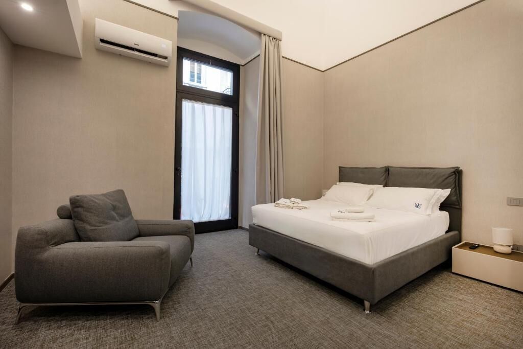 A modern hotel room with a bed, a sofa, an air conditioning unit, a window with curtains, and a nightstand with a lamp.