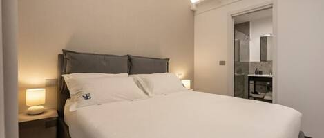 Una habitación de hotel con cama, mesita de noche, lámpara y baño visible a través de una puerta abierta.