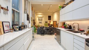 Ett smalt kök med vita skåp, en cykel och en matplats med en soffa och ett bord.