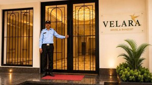 Entrada de un hotel con un hombre de pie frente a ella, un felpudo rojo y un letrero que dice 'VELARA HOTEL & BANQUET'.
