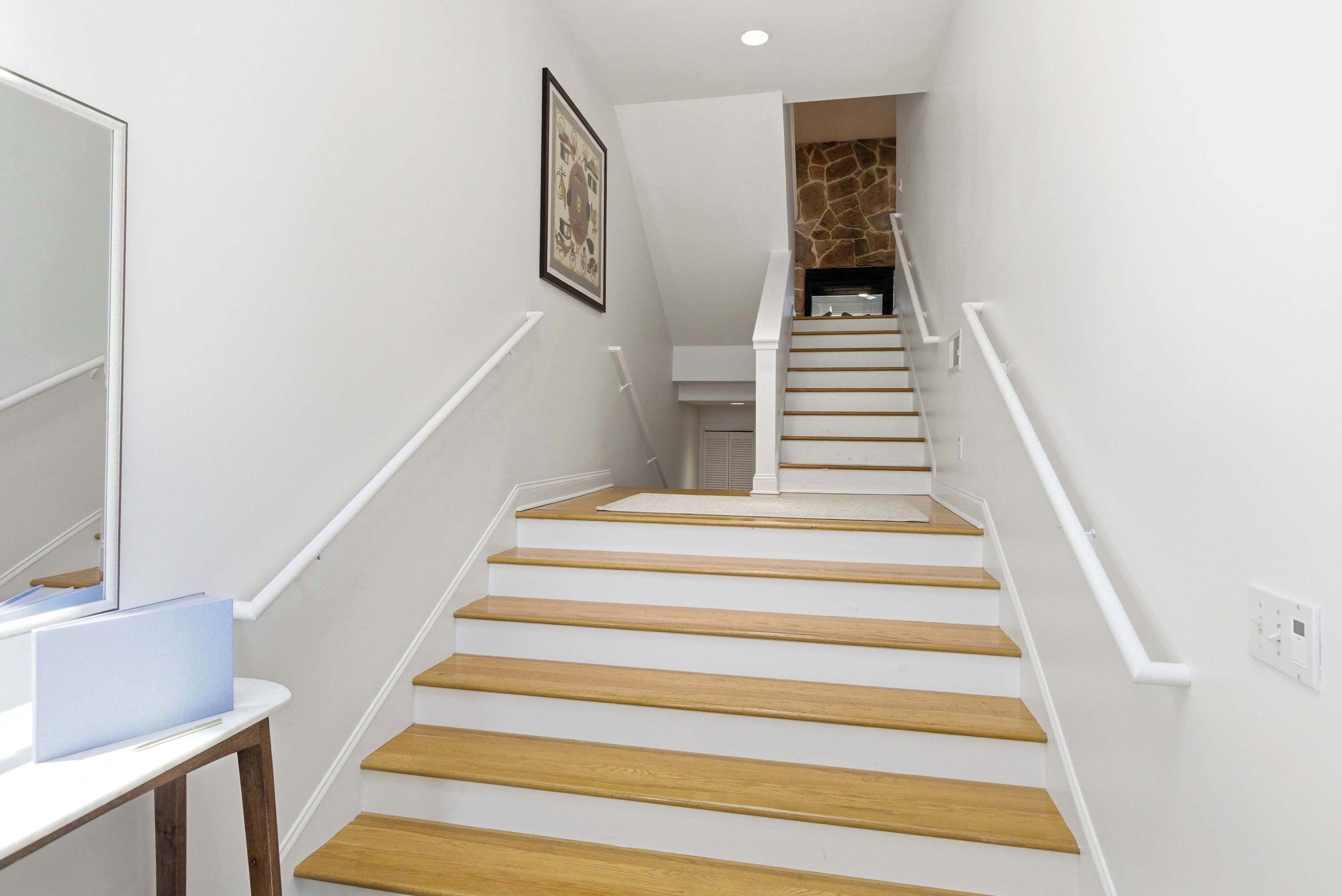 Un escalier avec des marches en bois et des rampes blanches, menant à un palier doté d’un mur en pierre et d’une cheminée.