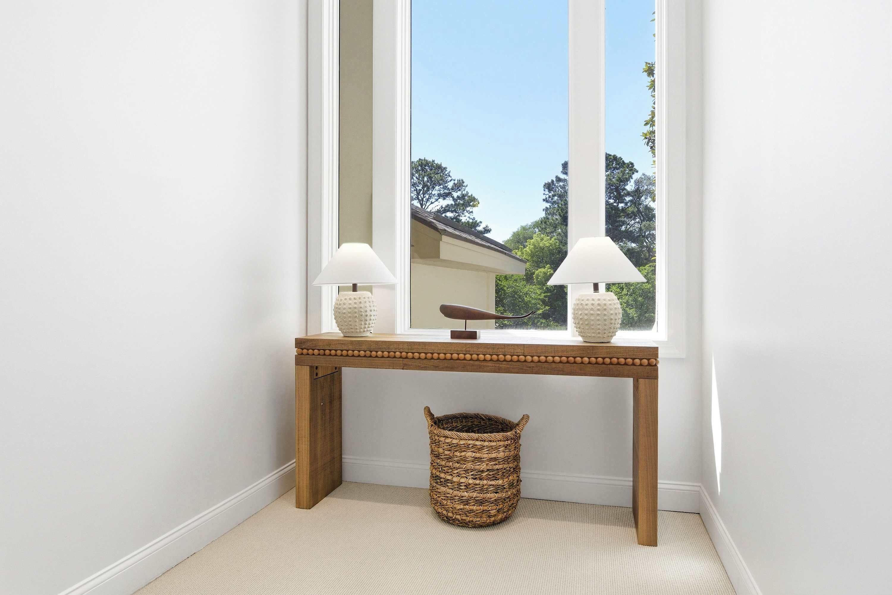 A corner with a wooden console table, two lamps, a woven basket, and a window with a view of trees.