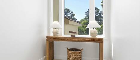 A corner with a wooden console table, two lamps, a woven basket, and a window with a view of trees.