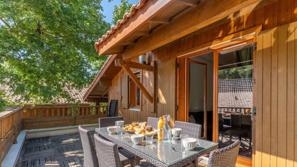 A wooden deck with wicker furniture, a glass table, and a door leading to a covered area. - Chic Family Flat With Heated Pool (Lacanau)