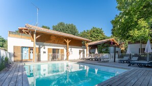 A swimming pool with wooden deck, a covered area with lounge chairs, and a building with a wooden facade. - Chic Family Flat With Heated Pool (Lacanau)