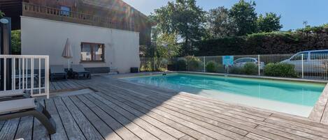 A wooden deck with a swimming pool, a covered seating area, and a fenced garden.
