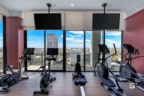A modern gym with various cardio machines, large windows offering city views, and mounted TVs. - Elite 1BR in Miami Worldcenter (Miami)