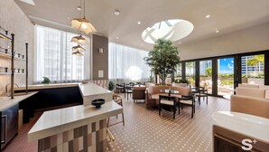 A modern kitchen with a central island, dining area with sofas, and a large window with a view of buildings. - Luxe 1-BR at Miami Worldcenter | Skyline (Miami)