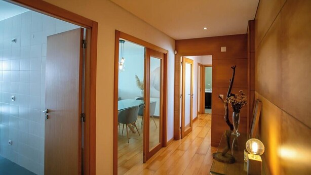 Um corredor com portas de madeira, uma sala de jantar com cadeiras e uma mesa, e um vaso decorativo com flores.