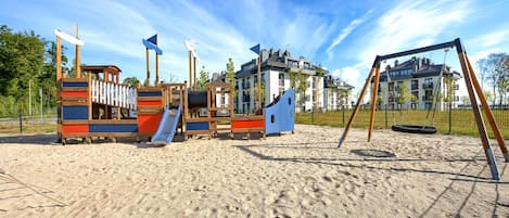 Ein Spielplatz mit Rutschen und Schaukeln auf Sandboden, umgeben von Gebäuden.