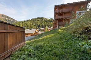 Un bâtiment en bois doté d’un balcon, entouré de verdure et adossé à une colline boisée.