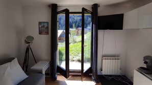 A room with a bed, a desk, a chair, a tripod, a television, and a view of a mountainous landscape through an open door. - Studio Apartment 'Le Refuge Douillet' with Mountain View and Private Terrace (La Plagne Tarentaise)