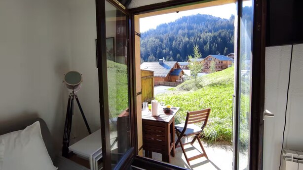 A room with a sliding glass door leading to a balcony with a table and chairs, a telescope, and a view of a forested hillside. - Studio Apartment 'Le Refuge Douillet' with Mountain View and Private Terrace (La Plagne Tarentaise)