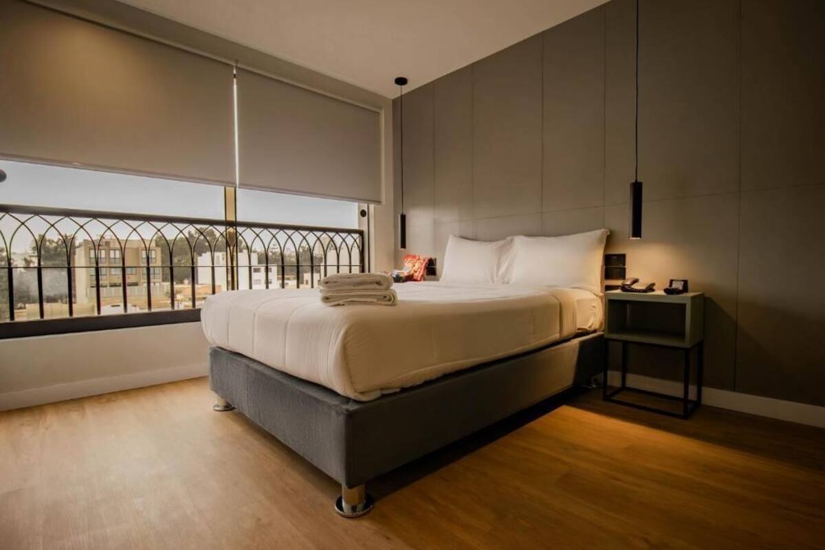 Un dormitorio moderno con una cama grande, una mesita de noche, una lámpara de pared y vistas a través de una ventana con barandilla de metal.