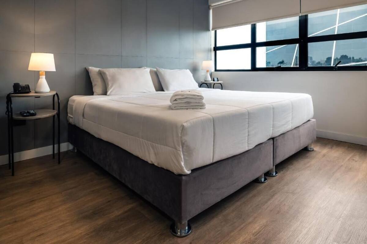 Habitación de hotel moderna con una cama grande, dos mesitas de noche, una lámpara y vistas a la ciudad a través de amplios ventanales.