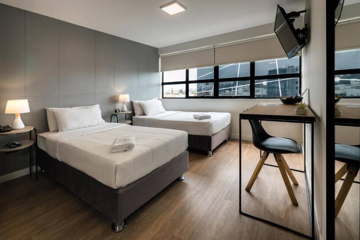 Una habitación de hotel moderna con dos camas, un escritorio, una silla, un televisor y una ventana con vista a la ciudad.