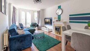 A living room with a blue sofa, a round coffee table, a fireplace, and a dining area with chairs. - Spacious Apt Stay - Near City Centre & Tram -Walk to Leith (Edinburgh)