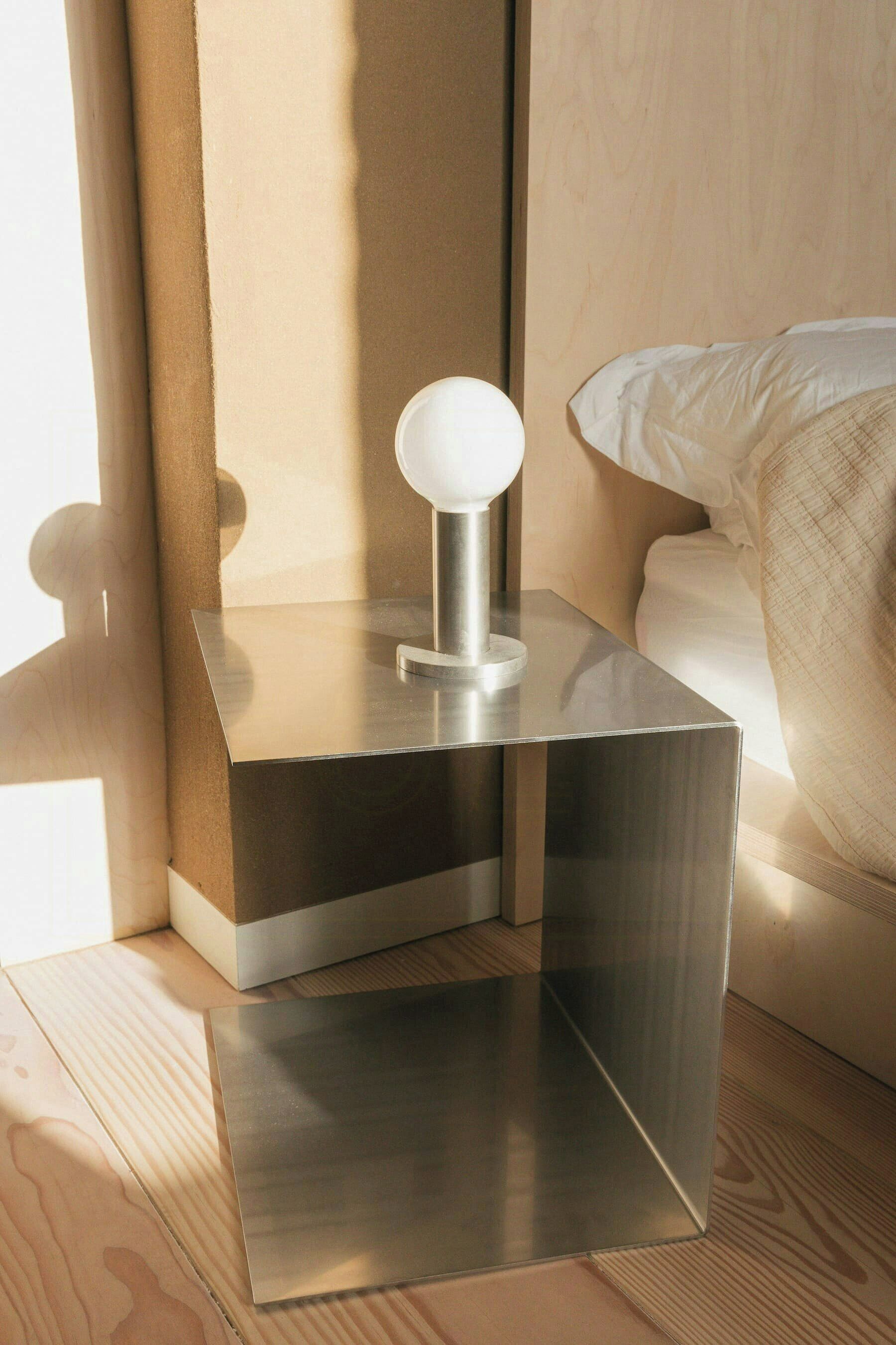 A modern bedside table with a metallic finish and a spherical object on top, positioned next to a bed with white bedding.