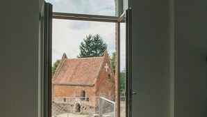 A room with a wooden floor, a sofa, and a large arched window leading to an outdoor area with a brick building. - Renovated commandery on the Elbe with sauna (Werben)