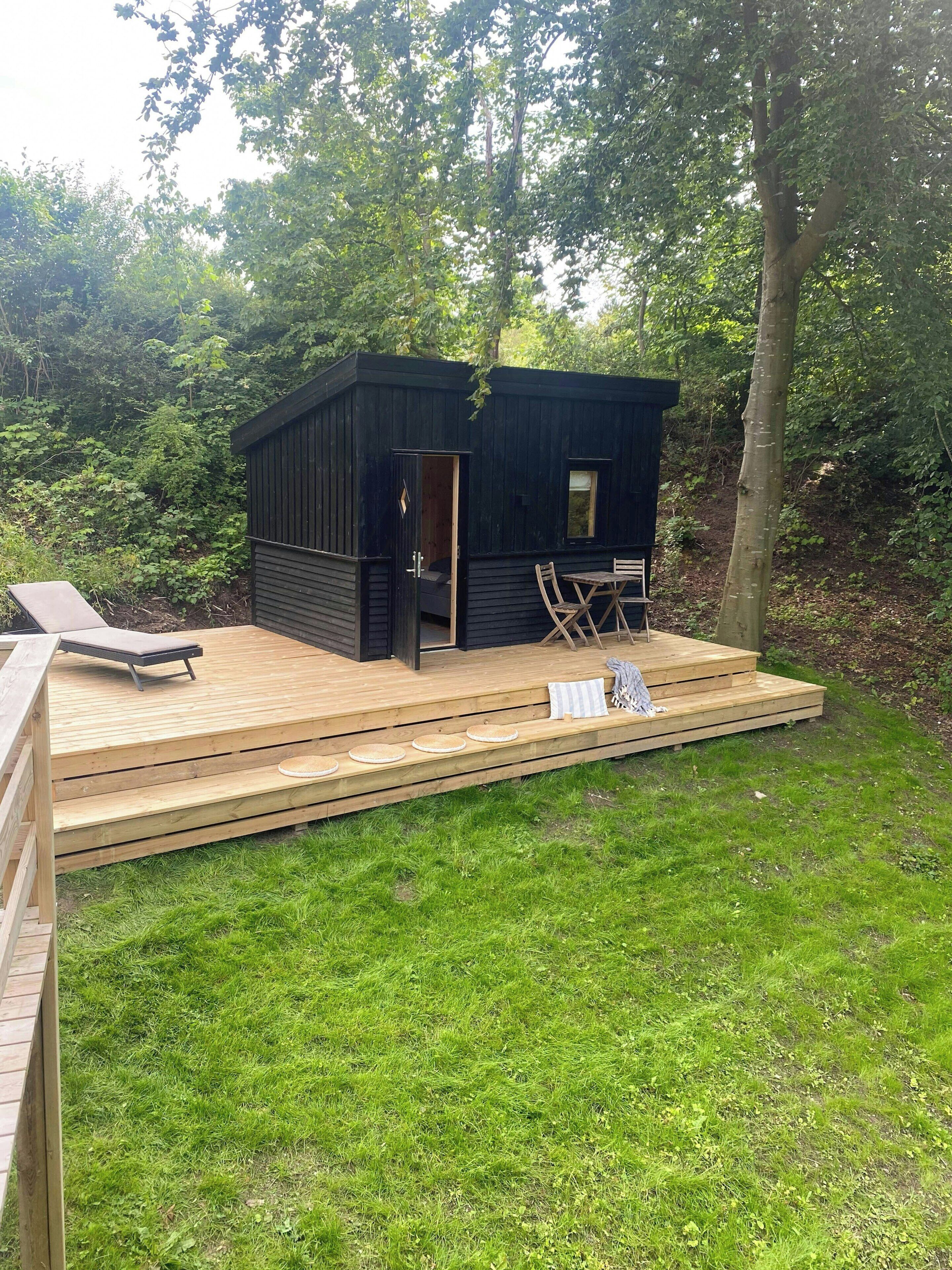 A small wooden cabin with a deck, a lounge chair, and a table.