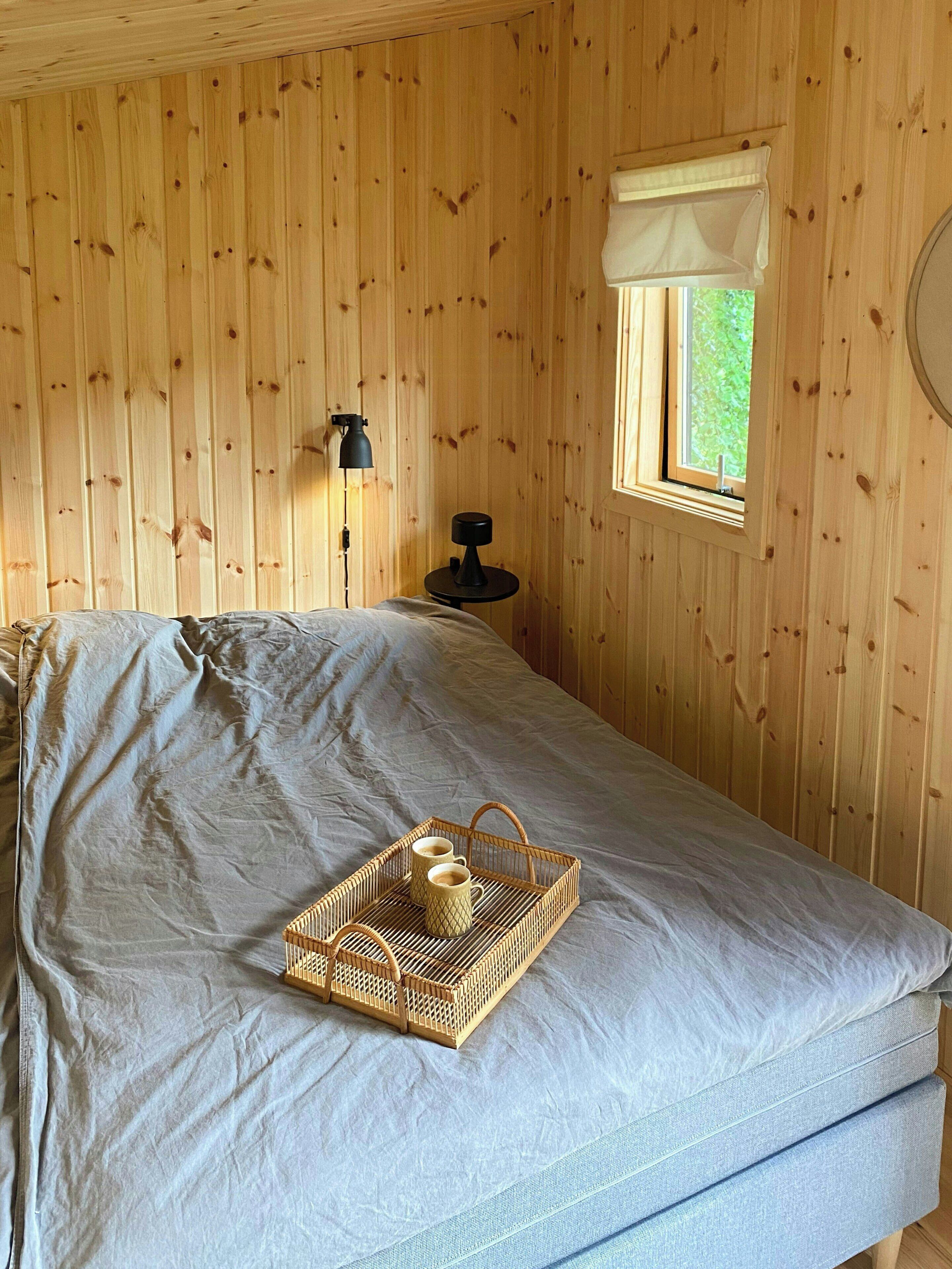 A room with wooden walls, a bed with a grey mattress, a bedside table with a lamp, and a window with a view of greenery.