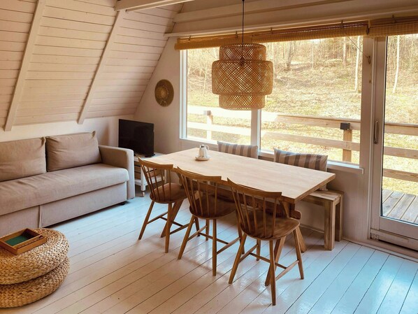 Une pièce confortable avec un canapé, une table à manger et des chaises, un téléviseur et une grande fenêtre donnant sur les arbres.