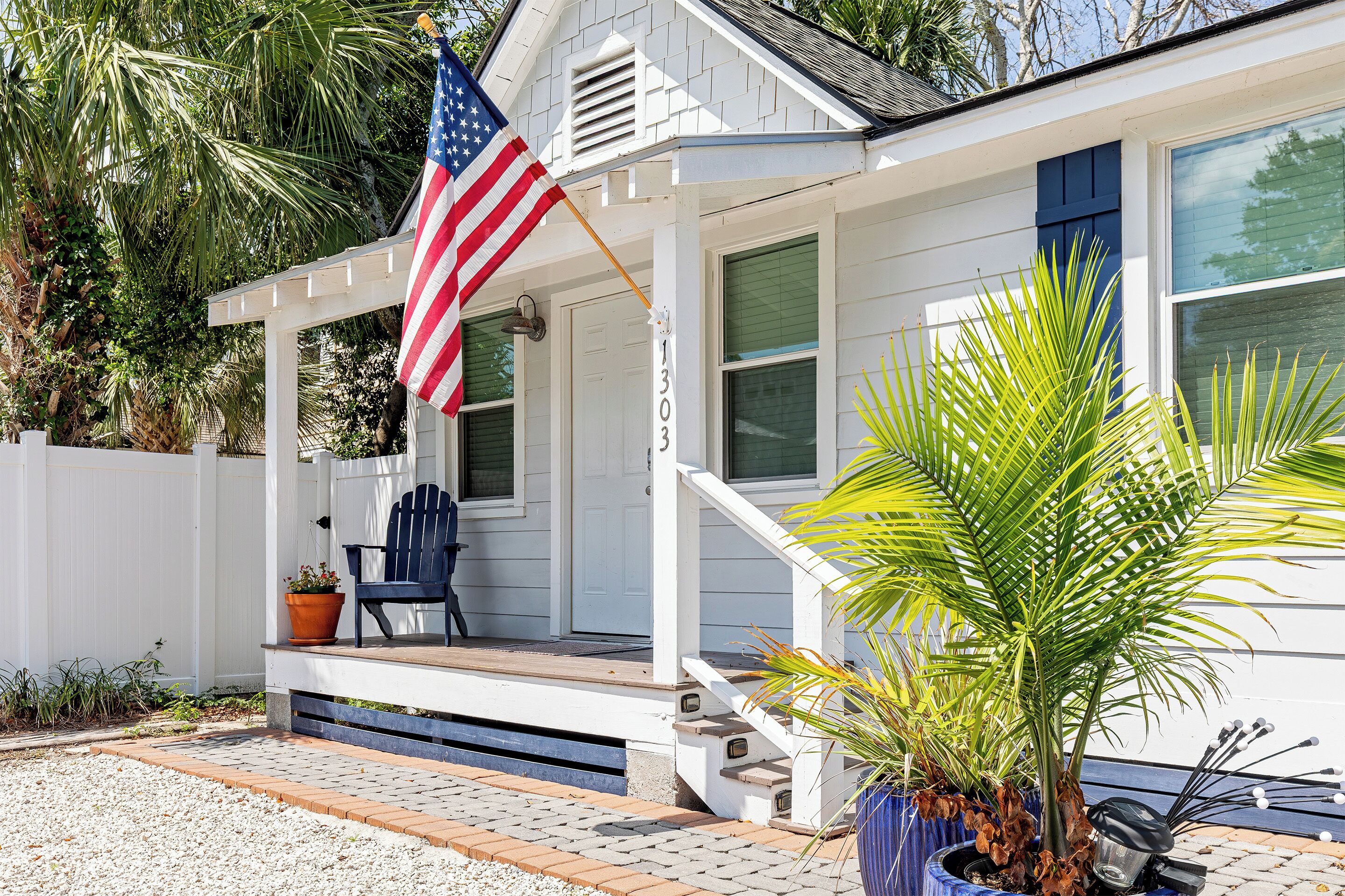 Et hvidt hus med veranda, en blå stol, en potteplante og et amerikansk flag.
