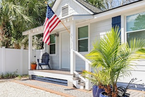 A white house with a porch, a blue chair, a potted plant, and an American flag. - Renovated, Quick Walk to Beach + Restaurants by Tybee Cottages (Tybee Island)