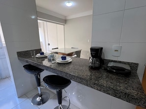 A modern kitchen with granite countertops, two black bar stools, a coffee maker, and a dining area with a table and chairs. - LK Santo Antônio 6  (Belo Horizonte)