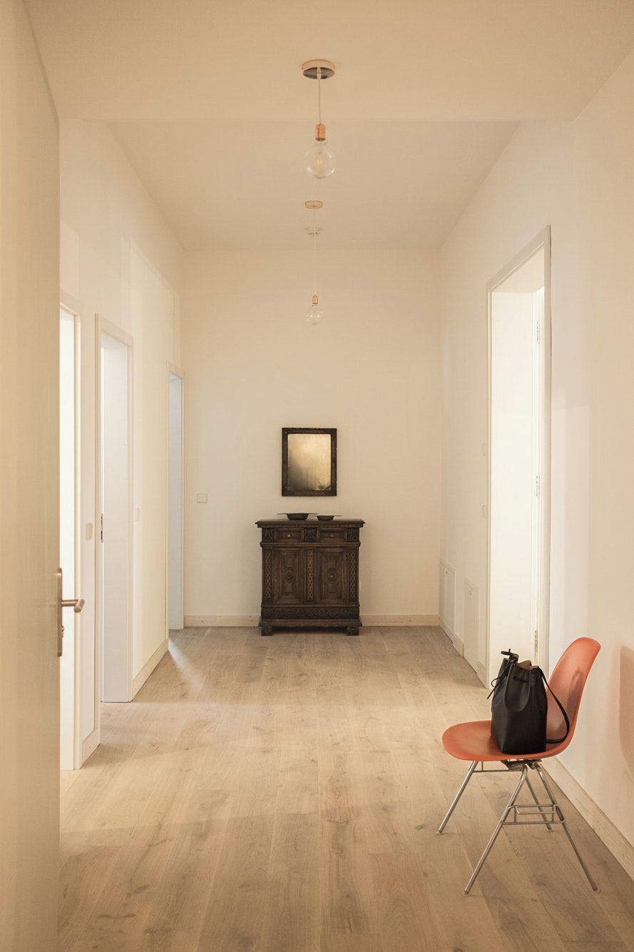 A hallway with a wooden floor, a dark wooden cabinet, a framed picture on the wall, and a red chair with a bag on it.