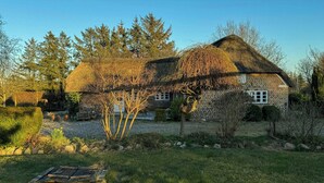 Una casa de piedra tradicional con tejado de paja, rodeada de un jardín cuidado y árboles.