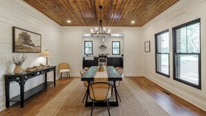 A dining area with a wooden ceiling, a chandelier, a long table, and a kitchen in the background.