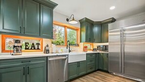 A kitchen with green cabinets, a stainless steel refrigerator, a dishwasher, and a window with a view of greenery.