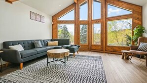 A modern living room with a dark grey sectional sofa, a round coffee table, and a patterned rug. Large windows provide a view of trees and a clear sky.