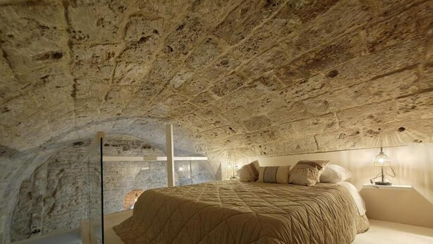 A bedroom with a stone ceiling, a bed with a quilt, a nightstand, and a lamp. - DOMUS GABRIEL (Giovinazzo)