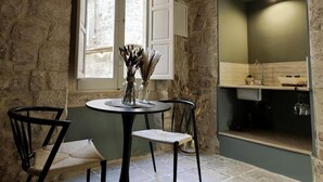 A stone-walled room with a round table, two chairs, a window with white shutters, and a kitchen area with a sink and countertop. - DOMUS GABRIEL (Giovinazzo)