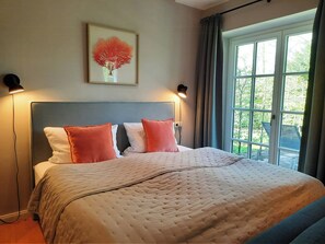 Ein Schlafzimmer mit einem Bett, zwei Nachttischlampen, einem Fenster mit Vorhängen und einem gerahmten Kunstwerk an der Wand.