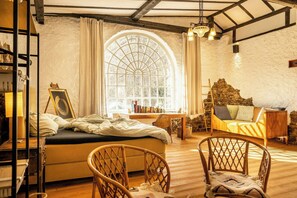 A cozy room with a bed, a wooden chair, a sofa, a large arched window, and a stone wall. - Immerse yourself into the magic of this studio and discover your creative side (Kall)